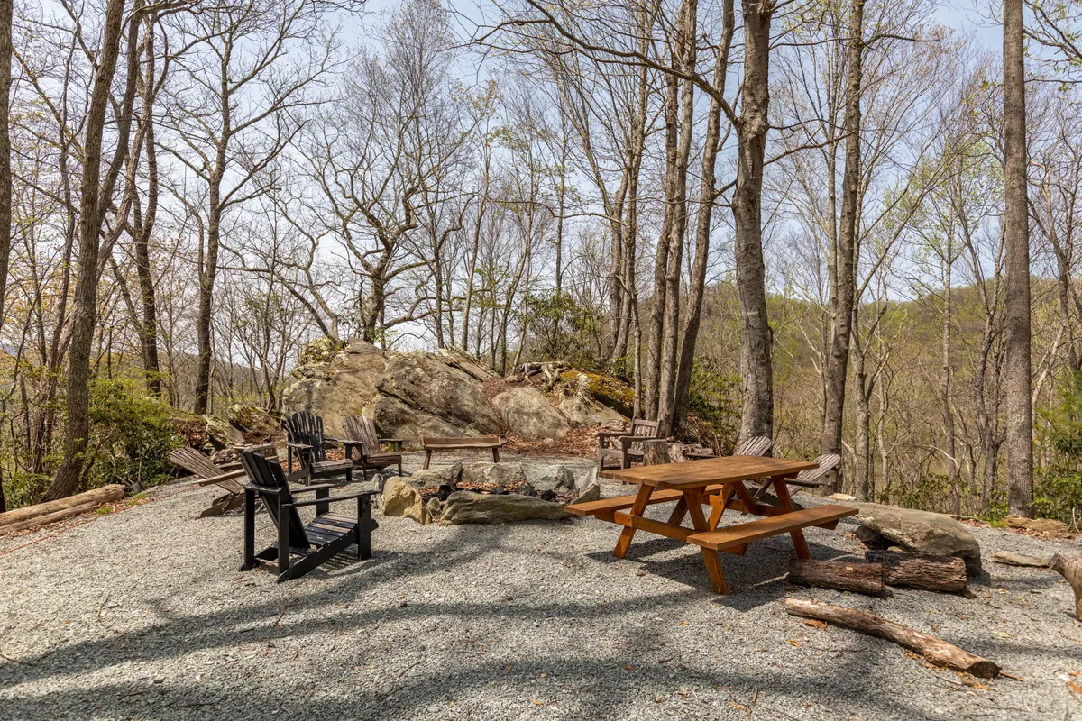 The image showcases a welcoming outdoor area featuring a fire pit surrounded by Adirondack chairs, a wooden picnic table, and natural rock formations. The space is set against a backdrop of rolling hills and natural greenery, perfect for outdoor gatherings and family activities under the open sky.