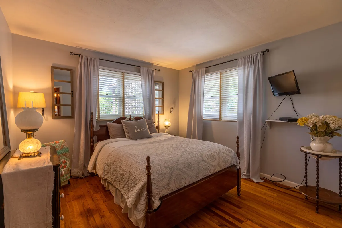 A cozy bedroom with a plush bed dressed in inviting linens sits by a window, accompanied by a stylish lamp and decorative pillows. Soft curtains frame the view, and a TV offers additional comfort and entertainment, capturing the restful ambiance ideal for unwinding after a day of adventure.