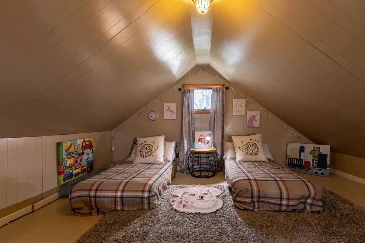A cozy interior bedroom featuring two twin beds with plaid bedding, a round rug with a dog design, natural light streaming through a window, and charming wall art. The playful rug and open space make it clear pets are welcome inside too.