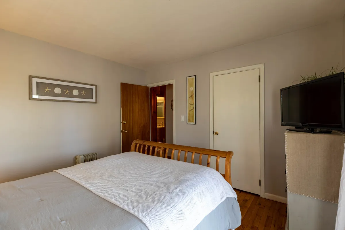 A peaceful bedroom scene with a neatly made bed, wall art, and a television, all illuminated by soft, natural light. The space feels tranquil and secluded, reflecting a restful escape within the cottage.