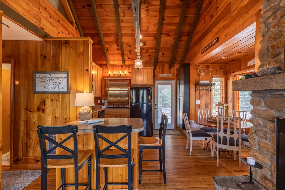 An inviting angle showing the seamless flow between the great room, dining area, and kitchen. The wooden beams and stone fireplace anchor the space while the kitchen island and dining table are clearly visible, showcasing areas for meal prep and shared dining.
