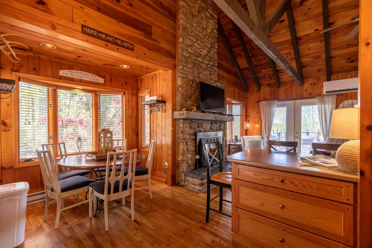 A warmly lit living and dining area featuring a wooden dining table next to a stone fireplace, with large windows revealing hints of the surrounding nature. The cozy setting is accented by natural wood cabinetry and decorative antlers, suggesting a rustic mountain retreat ideal for intimate moments.
