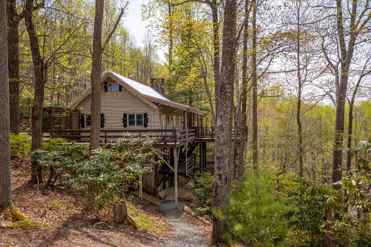 The hero image showcases a charming mountain cabin nestled among lush trees, with a welcoming pathway leading up to the front door. The scene is tranquil, emphasizing the cabin's seclusion and natural setting. The deck offers views ideal for enjoying sunset skies or a morning coffee, while hints of an outdoor fire pit area invite thoughts of cozy evenings under the stars.