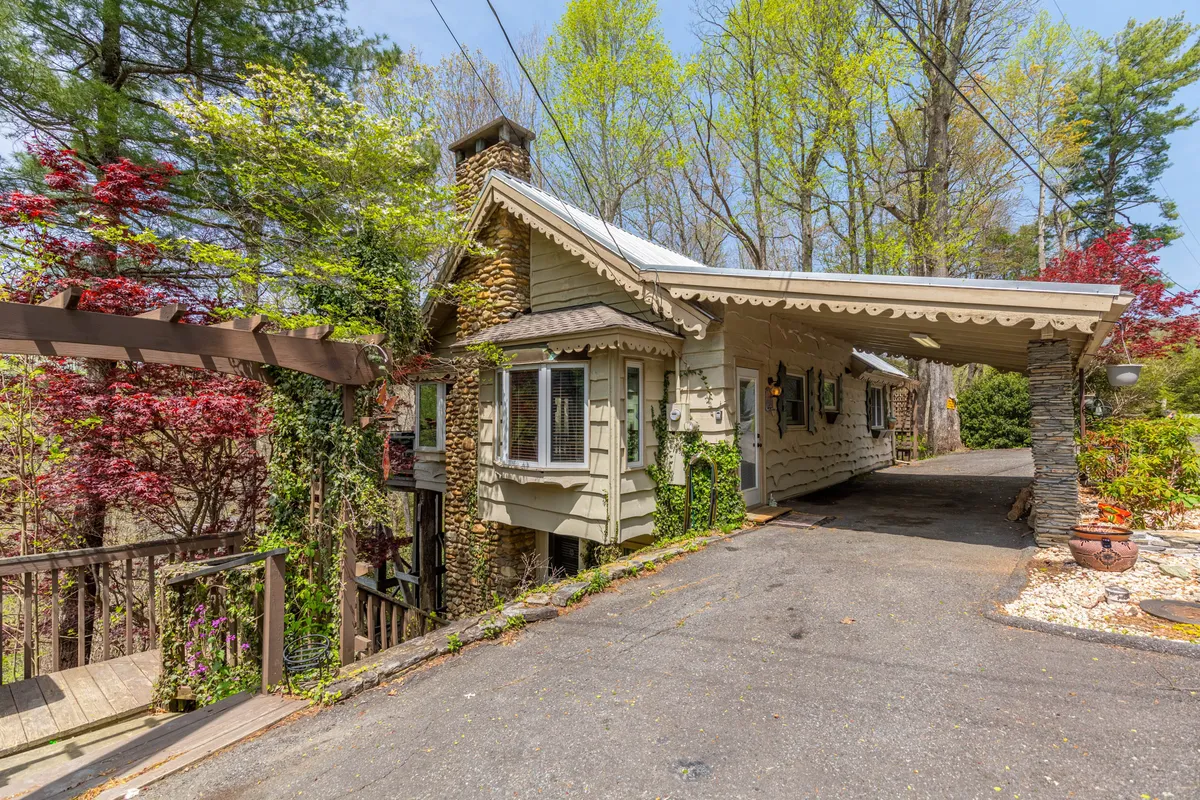 A wide view of the cottage exterior highlights the textured stone chimney, wrap-around wooden deck, lush greenery, and vibrant foliage. The deck is prepared for social gatherings, offering beautiful sunset views and a welcoming atmosphere that bridges the indoor and outdoor experience.