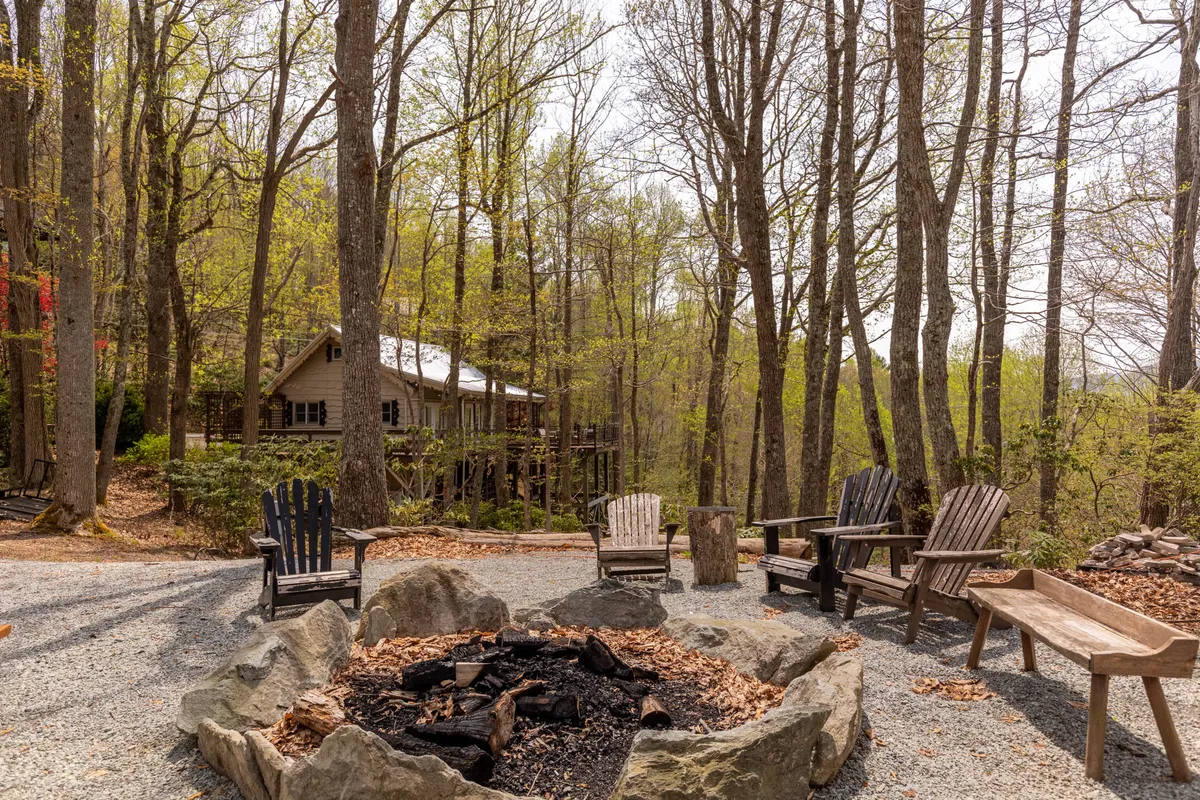 The fire pit area is shown with sturdy wooden chairs arranged around a blazing fire, surrounded by dense trees and a dramatic boulder outcropping. The cottage is visible in the background, grounding the scene in its mountain setting and showcasing the unique features of the property.