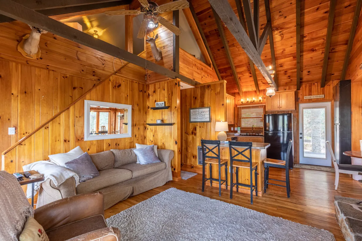 This image shows an open-plan living area with wooden beams, a comfortable sofa, a dining area, kitchen appliances, and large windows that bring in plenty of natural light. The space feels warm and inviting, with a clear visual connection to the outdoors, hinting at the mountain surroundings and the activities available beyond the cottage walls.