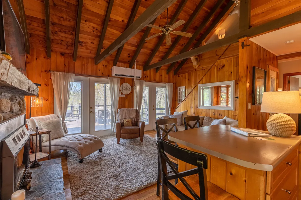 A spacious great room with vaulted wooden ceilings and prominent exposed beams. A grand stone fireplace serves as the focal point, surrounded by plush couches and a cozy seating area. Large windows fill the space with natural light, and the adjacent dining table invites group meals. The view through the windows hints at the tranquil deck outside.