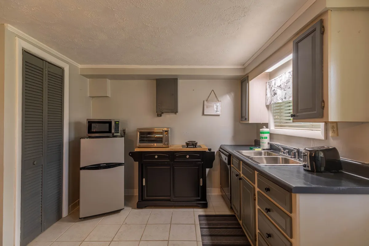 A compact and functional kitchenette area featuring a kitchen sink, microwave, stove, refrigerator, and a curtained window offering soft natural light. The setup is ideal for light meal preparation and quick snacks, aligning with the suite's amenities.